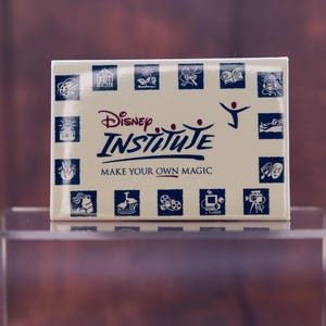 May include: A white box with blue and red accents. The box has the text "Disney Institute" and "Make Your Own Magic" on it. The box is surrounded by small blue squares with various images inside.