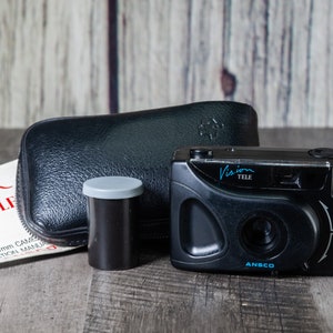 May include: A black Vision Tele camera with a tele-wide lens, a black leather camera case, and a roll of film. The camera has a built-in flash and a blue inscription that reads "Vision Tele" and "Ansco DX".