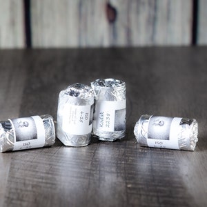 May include: Four rolls of film wrapped in silver foil, each with a white label. One label reads "Kodak 2238", while others show "ISO" and other details. The film is arranged on a wooden surface.