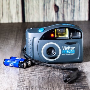 May include: A gray Vivitar EZ Motor Date-A-Print camera with a blue button and a black strap. Two blue AA batteries are included.