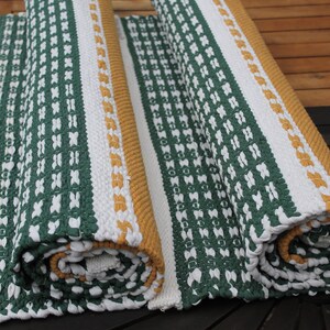 May include: Two rolled-up, woven rugs with a green and white pattern. One rug is partially unrolled, revealing a white border and a mustard-colored edge. The rugs are made of a thick, textured material.