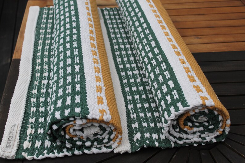 May include: Two rolled-up, woven rugs with a green and white pattern. One rug is partially unrolled, revealing a white border and a mustard-colored edge. The rugs are made of a thick, textured material.
