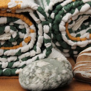 May include: Rolled green, white, and yellow patterned towels with a textured design. Two smooth stones in shades of gray and brown are in the foreground. The towels are tightly rolled, showcasing the pattern.