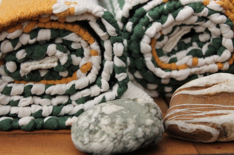 May include: Rolled green, white, and yellow patterned towels with a textured design. Two smooth stones in shades of gray and brown are in the foreground. The towels are tightly rolled, showcasing the pattern.
