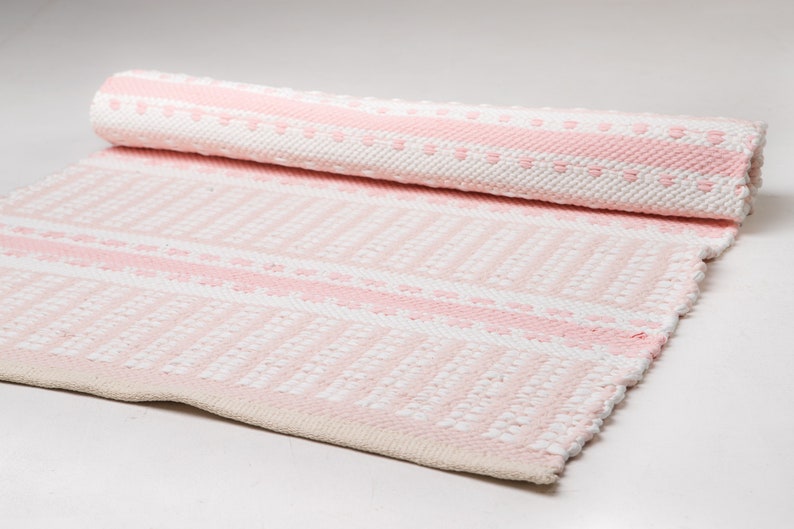 Puede incluir: Una alfombra tejida a rayas rosa y blanca con un patr&oacute;n texturizado. La alfombra est&aacute; enrollada y los bordes son visibles.