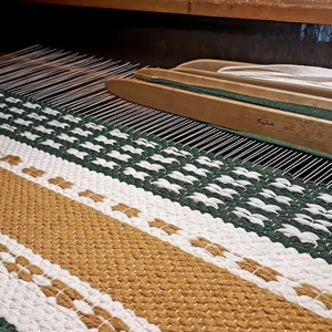 May include: A woven textile in shades of gold, white, and green is displayed on a loom. The text "Herman Tekstiil" is visible. The textile has a striped pattern with a textured appearance, showcasing the craftsmanship of the weaving process.