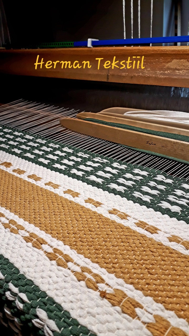 May include: A woven textile in shades of gold, white, and green is displayed on a loom. The text "Herman Tekstiil" is visible. The textile has a striped pattern with a textured appearance, showcasing the craftsmanship of the weaving process.