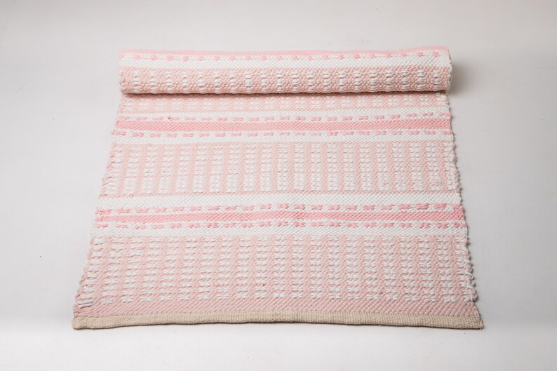 Puede incluir: Una alfombra tejida rosa y blanca con un patr&oacute;n geom&eacute;trico. La alfombra est&aacute; enrollada en la parte superior.