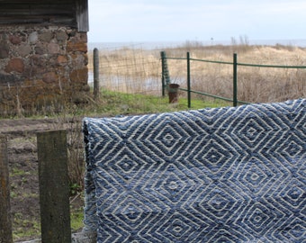 Op de afbeelding: Een blauw en wit geweven tapijt met een ruitpatroon hangt aan een houten hek. Het tapijt is gemaakt van natuurlijke vezels en heeft een rustieke uitstraling.