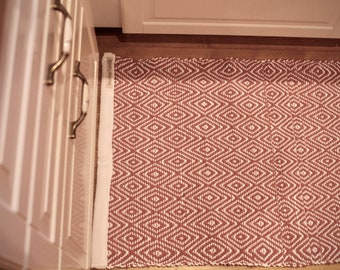 Op de afbeelding: Een rood en wit ruitjespatroon tapijt met een witte rand. Het tapijt ligt voor een witte kast met gouden handvatten.