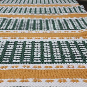 May include: A woven table runner with a repeating pattern of green, white, and gold stripes. The runner is made of a textured material and is suitable for a dining table or other surface.