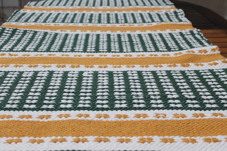 May include: A woven table runner with a repeating pattern of green, white, and gold stripes. The runner is made of a textured material and is suitable for a dining table or other surface.