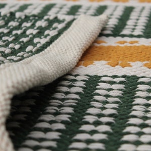 May include: Close-up of a woven rug with a green, white, and yellow pattern. The rug's edge is folded over, revealing a thick, off-white border. The design features vertical green stripes and horizontal yellow stripes.