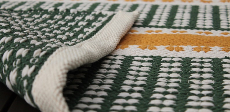 May include: Close-up of a woven rug with a green, white, and yellow pattern. The rug's edge is folded over, revealing a thick, off-white border. The design features vertical green stripes and horizontal yellow stripes.