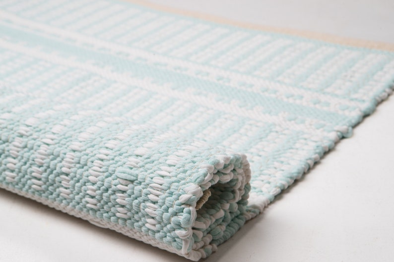 Puede incluir: Una alfombra tejida de color azul claro y blanco con un patr&oacute;n geom&eacute;trico. La alfombra est&aacute; enrollada y la textura del tejido es visible.