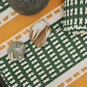 May include: A woven textile with a green, white, and yellow geometric pattern. Two stones and a woven basket are on the textile. The textile has a striped design with alternating colors.