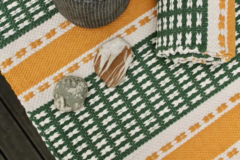 May include: A woven textile with a green, white, and yellow geometric pattern. Two stones and a woven basket are on the textile. The textile has a striped design with alternating colors.