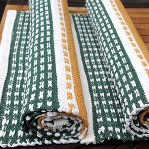 May include: Rolled-up green, white, and gold woven rugs. The rugs feature a repeating pattern of white crosses on a green background, with white and gold borders. The rugs are made of a thick, textured material and appear to be handmade.