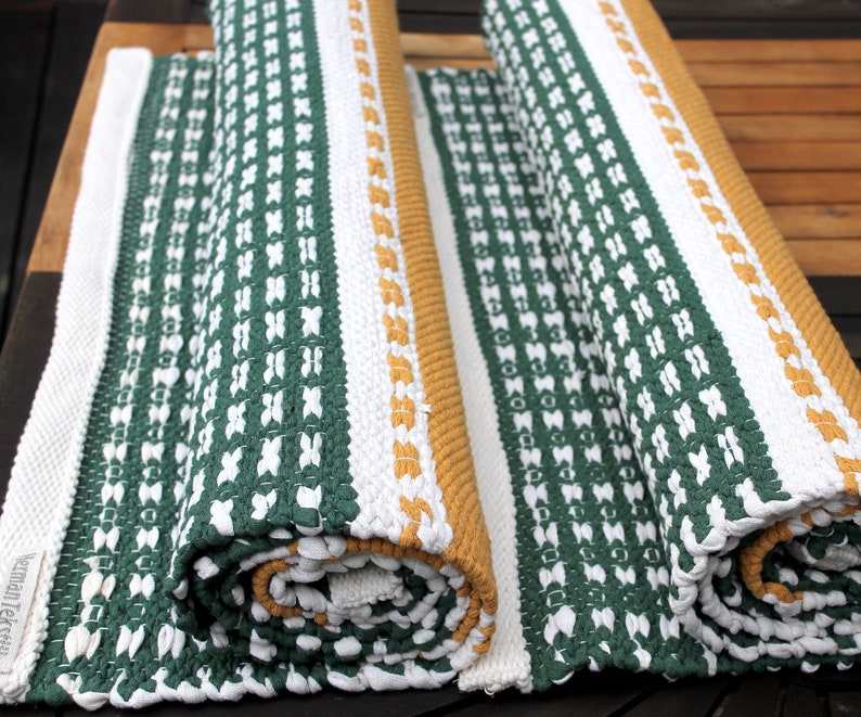 May include: Rolled-up green, white, and gold woven rugs. The rugs feature a repeating pattern of white crosses on a green background, with white and gold borders. The rugs are made of a thick, textured material and appear to be handmade.