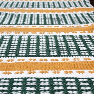 May include: A close-up of a woven rug with a repeating pattern of green, white, and gold horizontal stripes. The rug has a textured surface and is likely made of natural fibers. The design is geometric and the colors are earthy.