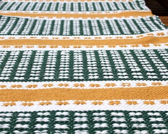 May include: A close-up of a woven rug with a repeating pattern of green, white, and gold horizontal stripes. The rug has a textured surface and is likely made of natural fibers. The design is geometric and the colors are earthy.