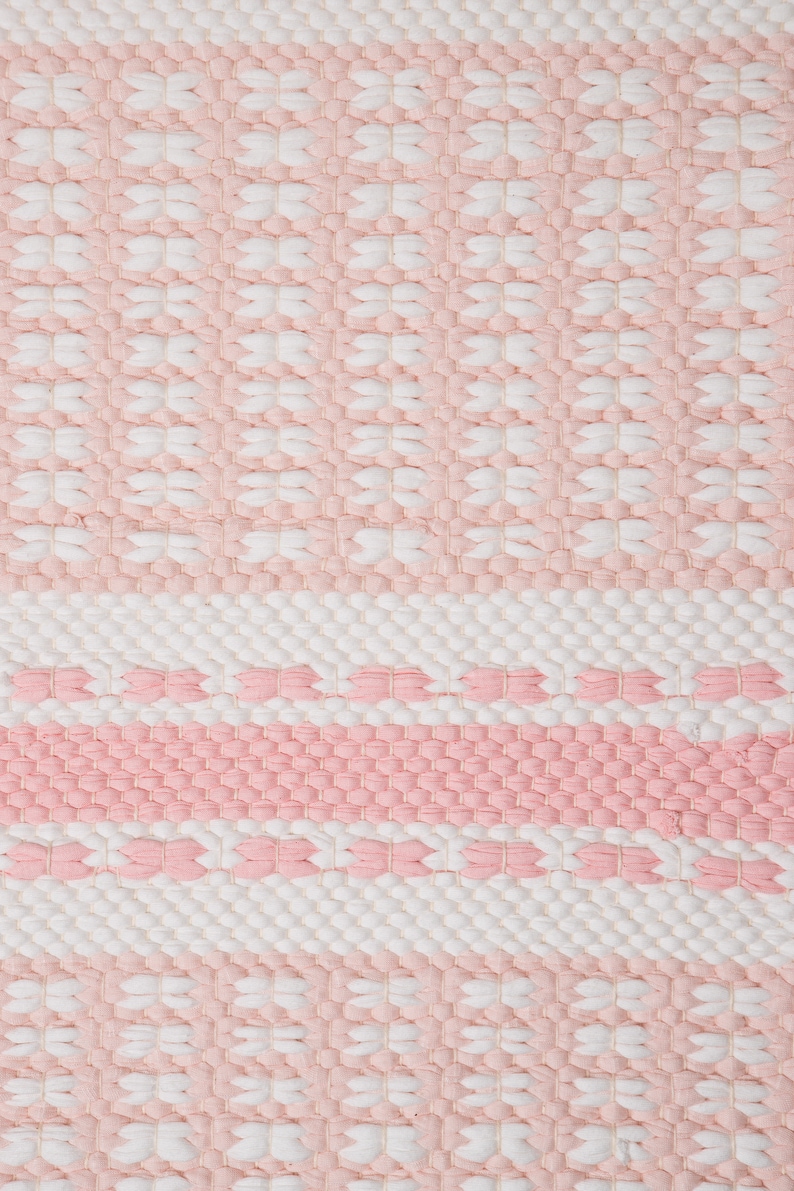 Puede incluir: Una alfombra tejida de color rosa y blanco con un patr&oacute;n geom&eacute;trico. La alfombra tiene una superficie texturizada y est&aacute; hecha de fibras naturales suaves.