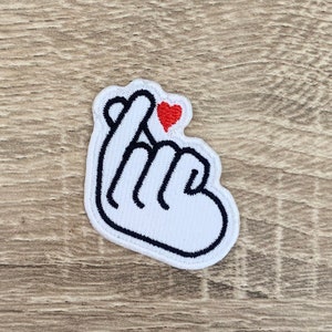 May include: White embroidered patch of a hand making the Korean heart sign with a small red heart on the index finger.