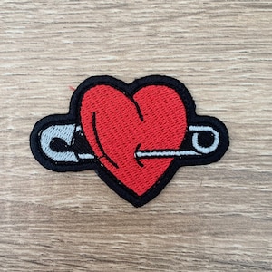 Puede incluir: Un corazón rojo con un imperdible atravesándolo. El corazón y el imperdible están bordados en un parche de tela.