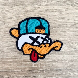 Op de afbeelding: Een geborduurde strijk-patch met een cartoon eend die een blauwe en witte baseballpet draagt met een 'X' over zijn ogen. De eend heeft een gele snavel en een rode tong die uitsteekt.