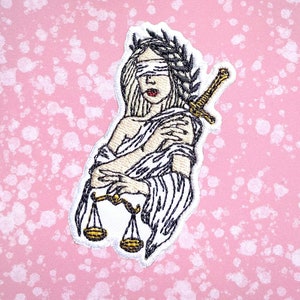 Può includere: Toppa ricamata da stirare con una figura bendata che tiene una bilancia e una spada. La figura indossa una corona di foglie e una veste fluida. La toppa è bianca con contorni neri e dettagli dorati.