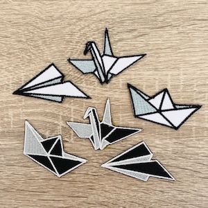 Może przedstawiać: Sześć naszywek do prasowania z motywami origami w kolorze czarnym i białym. Wzory obejmują łódkę, samolot i żurawia.
