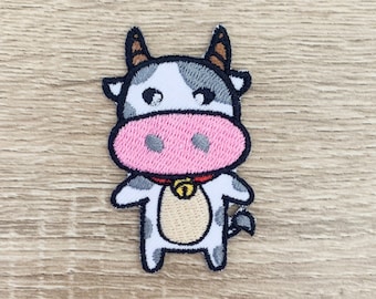 May include: A white and black cow with a pink nose and a red collar with a gold bell. The cow is standing on a wooden background.
