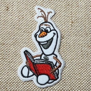 Könnte beinhalten: Gestickter Aufnäher mit Olaf aus Frozen, der ein Buch liest. Der Aufnäher hat einen weißen Umriss, Olafs Körper ist weiß, die Nase orange und die Haare braun. Das Buch ist rot mit schwarzem Text.