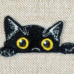 Könnte beinhalten: Gestickter schwarzer Katzenaufnäher mit großen gelben Augen und weißen Akzenten. Die Katze lugt über eine Kante, ihre Pfoten sind sichtbar. Der Aufnäher befindet sich auf einem beigen Stoffhintergrund.