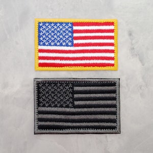 Può includere: Due toppe ricamate con la bandiera americana. La toppa superiore è rossa, bianca e blu con un bordo giallo. La toppa inferiore è nera e grigia.