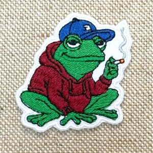 以下が含まれることがあります： 赤いパーカーと青い野球帽をかぶった緑色のカエルの刺繍パッチ。カエルはタバコを吸っています。パッチは白い縁取りで、ベージュ色の生地の背景にあります。