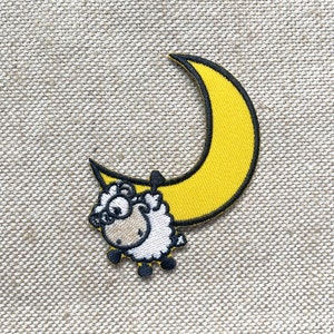 May include: Embroidered patch showing a cartoon sheep in glasses, suspended from a yellow crescent moon with a black border. The patch is set against a textured, neutral backdrop.