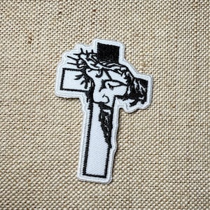Op de afbeelding: Witte geborduurde patch van een persoon op een kruis met een doornenkroon. De patch is omrand met zwart garen.