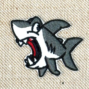 May include: Embroidered patch of a cartoon shark with an open mouth, revealing teeth and a red interior. The shark has a white body, grey fins, and a black outline. The patch is set against a beige fabric background.