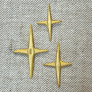 Puede incluir: Tres parches bordados dorados en forma de estrella. Cada estrella tiene cuatro puntas alargadas. Los parches están dispuestos sobre un fondo de tela gris claro texturizado. Las estrellas tienen un brillo metálico.