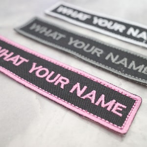 Può includere: Tre toppe di tessuto nero con cuciture rosa. Ogni toppa dice "WHAT YOUR NAME" in lettere bianche, grigie e rosa.