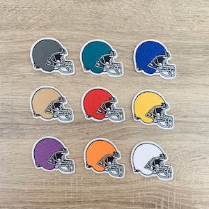 Op de afbeelding: Negen geborduurde voetbalhelm-patches in verschillende kleuren, waaronder groen, blauw, rood en geel. Elke patch heeft een gedetailleerd zilveren gezichtsmasker en een witte rand, gerangschikt op een licht houtnerfoppervlak.