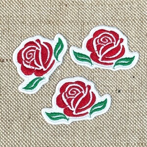 May include: Three embroidered rose patches. Each patch features a red rose with white detailing and green leaves, all outlined in white. The patches are arranged on a textured, tan background.