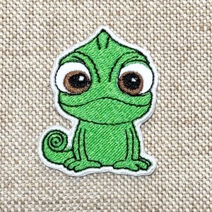May include: Embroidered patch featuring a green chameleon with large brown eyes and a white outline. The cartoon-style design has a curled tail and is set against a neutral background.