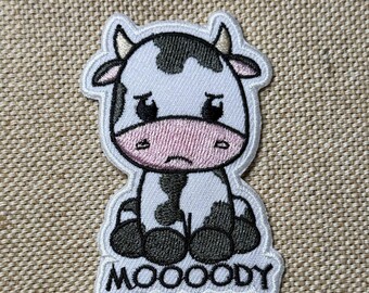 May include: White and black embroidered iron-on patch of a cartoon cow with a sad expression. The cow is sitting down with its legs crossed. The text "MOOOODY" is embroidered below the cow.