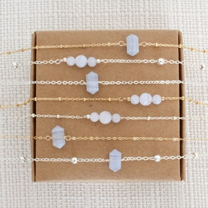 Peut inclure: Un ensemble de huit colliers délicats avec différentes chaînes en argent et en or et des accents de pierres précieuses bleu clair. Les colliers sont présentés sur une boîte en carton marron.