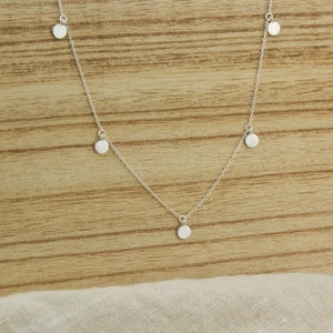 Puede incluir: Un delicado collar de plata con pequeños colgantes redondos en forma de disco. El collar tiene una fina cadena y múltiples colgantes espaciados uniformemente a lo largo de su longitud. Los colgantes son reflectantes y captan la luz, creando un brillo sutil.