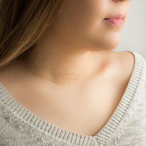 Puede incluir: Primer plano de una persona que lleva dos delicados collares de oro. Los collares son cadenas finas que se colocan cerca del cuello. La persona lleva un su&eacute;ter gris.