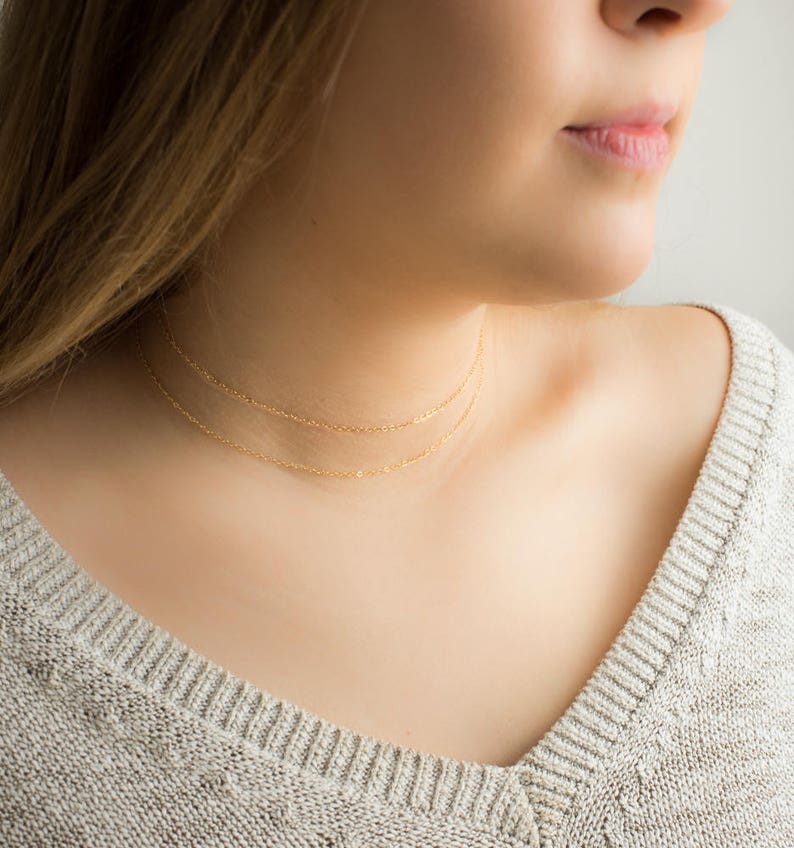 Puede incluir: Primer plano de una persona que lleva dos delicados collares de oro. Los collares son cadenas finas que se colocan cerca del cuello. La persona lleva un su&eacute;ter gris.
