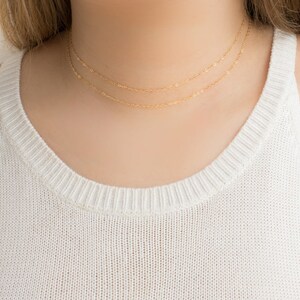 Puede incluir: Primer plano de una persona con una camiseta sin mangas blanca y dos delicados collares dorados. Los collares est&aacute;n superpuestos, uno ligeramente m&aacute;s largo que el otro. Las cadenas de oro son finas y elegantes.